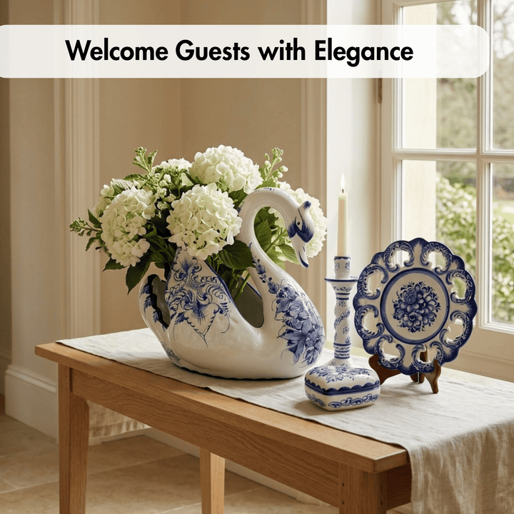 A white ceramic swan-shaped flower vase with blue floral patterns holds white hydrangeas on a wooden table. A matching blue-and-white plate, candlestick, and small box sit beside it near a window, with the text &