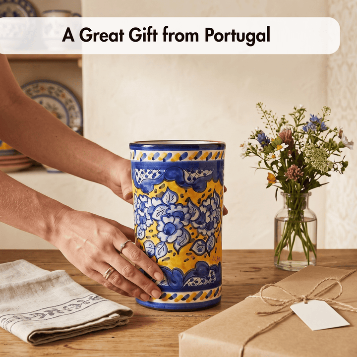 A person holds a tall ceramic canister with a yellow and blue floral pattern, set on a wooden table next to a wrapped gift box and a small vase of flowers. Text at the top reads &