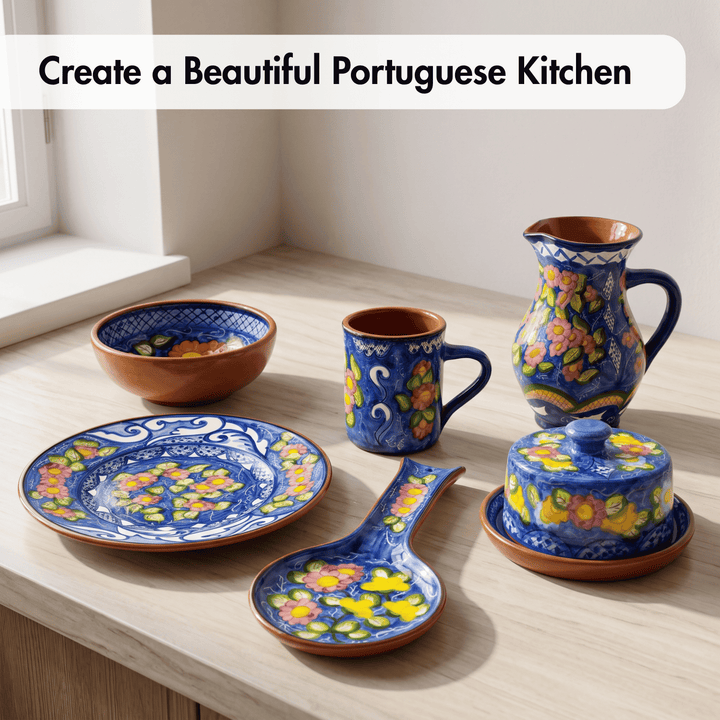 A set of hand-painted ceramic tableware displayed on a light wood countertop near a window. The set includes a plate, bowl, mug, pitcher, lidded dish, and spoon, all decorated with a vibrant blue base, yellow and pink floral motifs, and terracotta-colored rims.