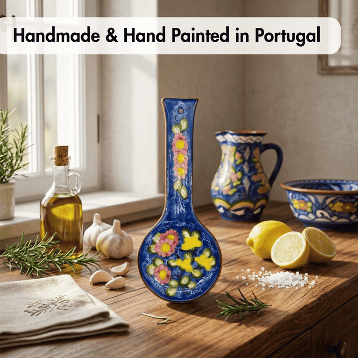 A colorful handmade ceramic vase with a long neck and floral hand-painted design stands on a wooden table in a kitchen setting, surrounded by lemons, garlic, rosemary, a bottle of olive oil, and other matching decorated ceramics. Text at the top reads &