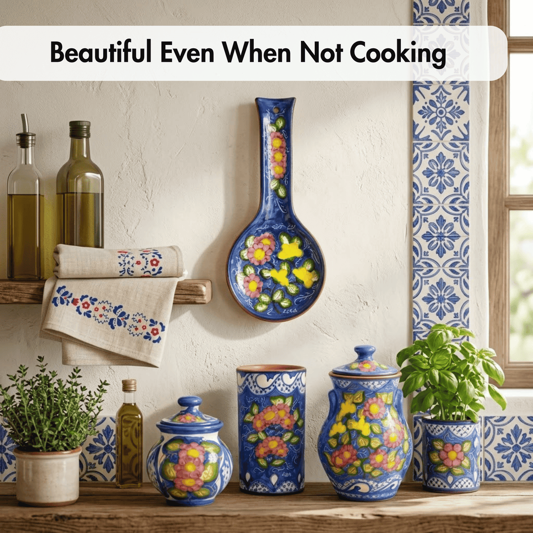 A styled kitchen scene with colorful ceramic canisters, a spoon rest, and a hanging bowl decorated with floral patterns in blue, yellow, and pink. A towel with matching embroidery hangs on a wooden shelf beside olive oil bottles, with potted plants and blue-and-white tile accents against a textured wall.