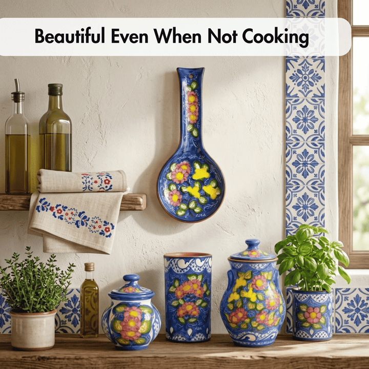 A styled kitchen scene with colorful ceramic canisters, a spoon rest, and a hanging bowl decorated with floral patterns in blue, yellow, and pink. A towel with matching embroidery hangs on a wooden shelf beside olive oil bottles, with potted plants and blue-and-white tile accents against a textured wall.