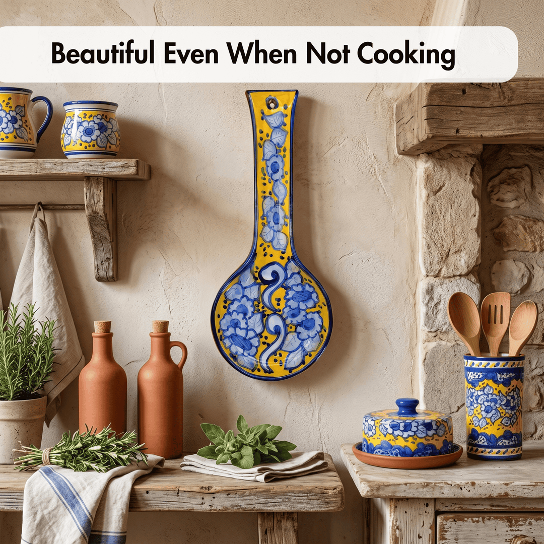 A rustic kitchen scene with a large decorative blue and yellow spoon-shaped wall plaque hanging on a beige wall. The scene includes shelves, terracotta bottles, herbs, a cloth towel, wooden utensils, and a lidded ceramic dish.