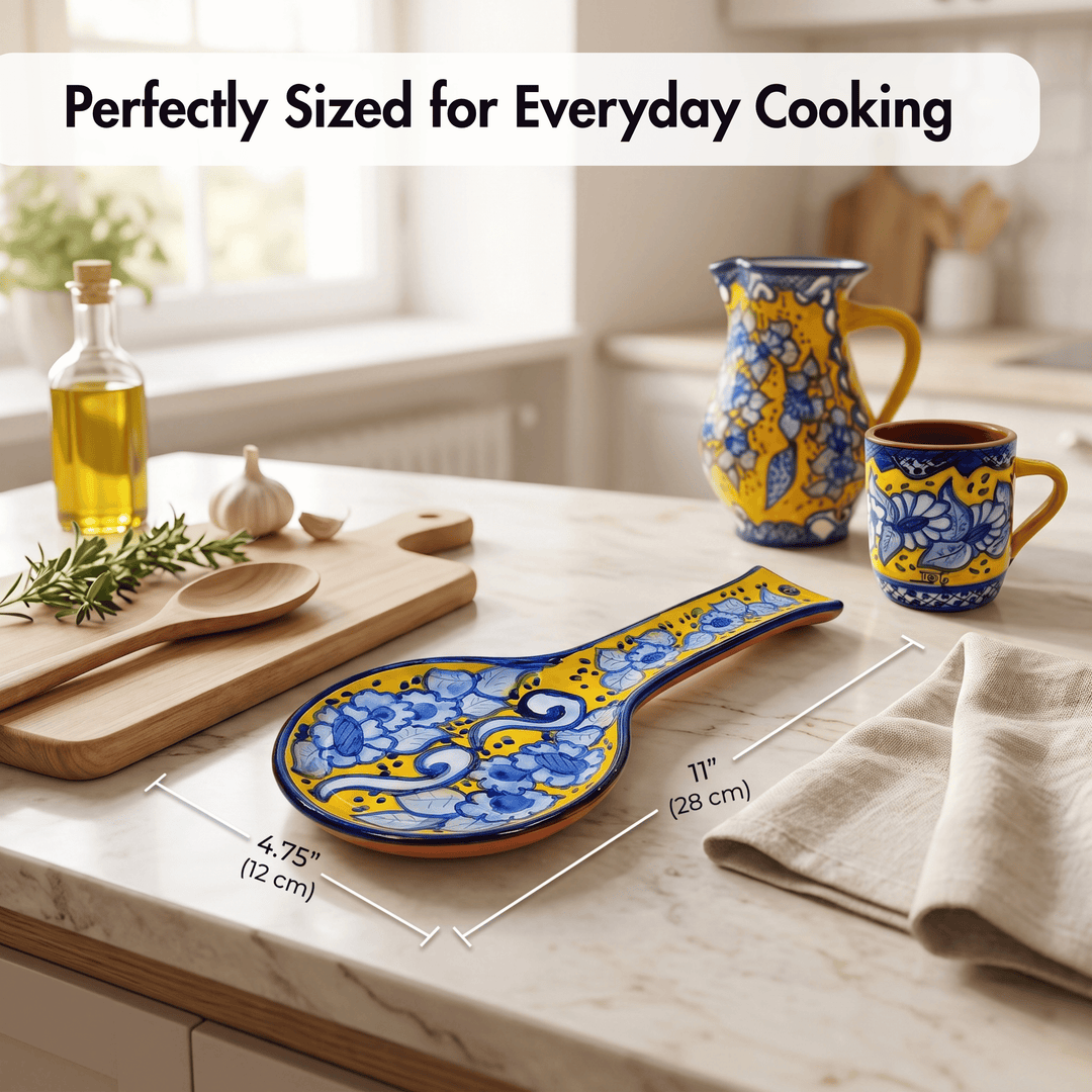 A ceramic spoon rest with a blue and yellow floral pattern is shown on a kitchen countertop, with dimension lines indicating its size. The scene includes matching yellow and blue cookware, a cutting board, garlic, olive oil, and a beige towel, with the text &