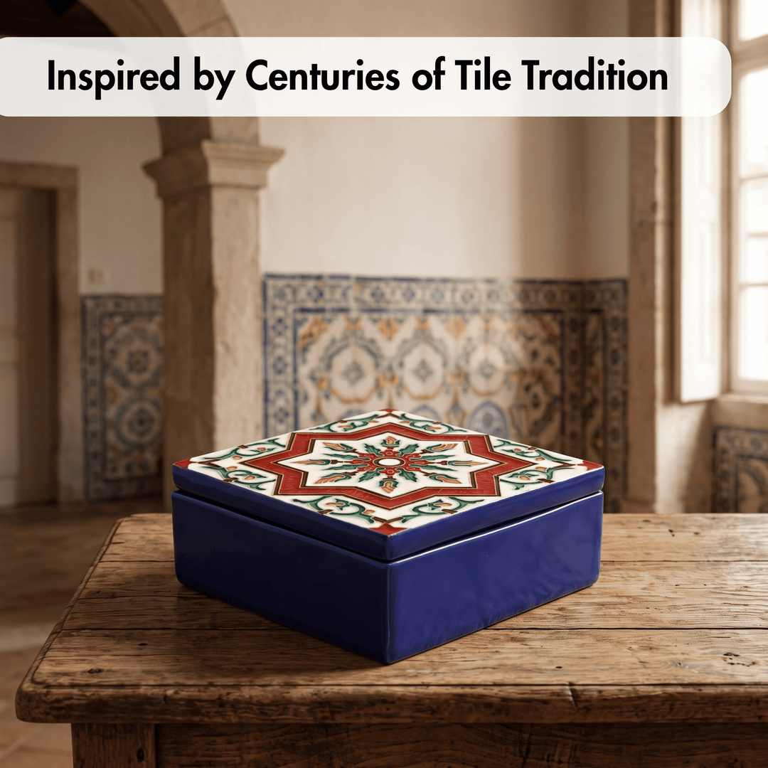 A square ceramic box with a lid, featuring a deep cobalt blue base and a decorative tile pattern on the lid in red, green, cream, and white. The box sits on a rustic wooden table in a room with tiled walls, with a text overlay at the top reading &