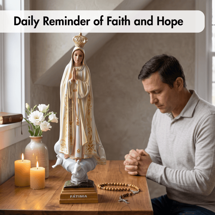 A man in a light gray long-sleeve shirt sits at a wooden table, praying with his hands clasped. On the table is a religious statue of Our Lady of Fatima wearing a white robe with gold embroidery, a wooden cross, and a rosary.