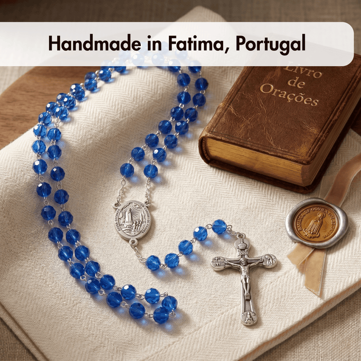 Blue rosary with silver crucifix and medal on a brown book, with text 'Handmade in Fatima, Portugal'.