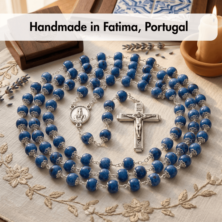 Blue rosary with silver crucifix on a decorative cloth with text 'Handmade in Fatima, Portugal'.