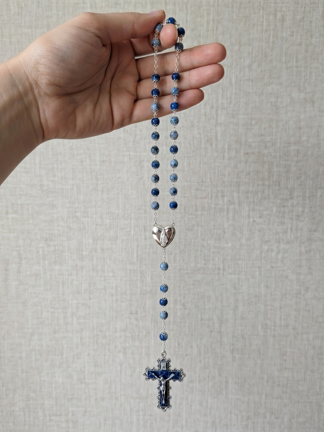 Hand holding a blue rosary with a silver heart charm and cross on a light gray background