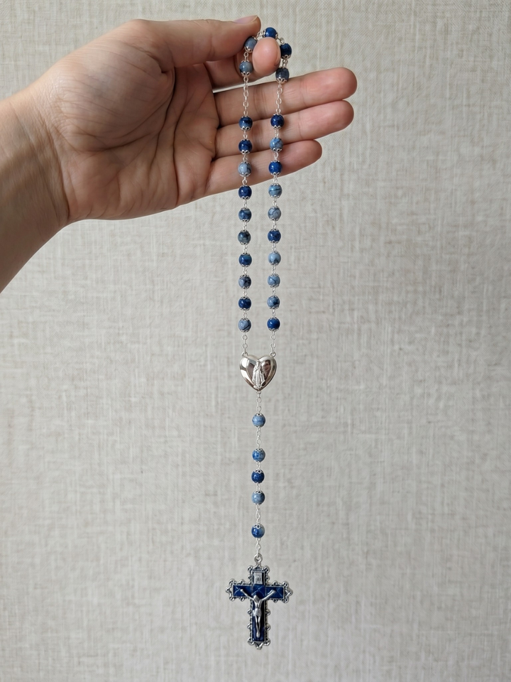 Hand holding a blue rosary with a silver heart charm and cross on a light gray background