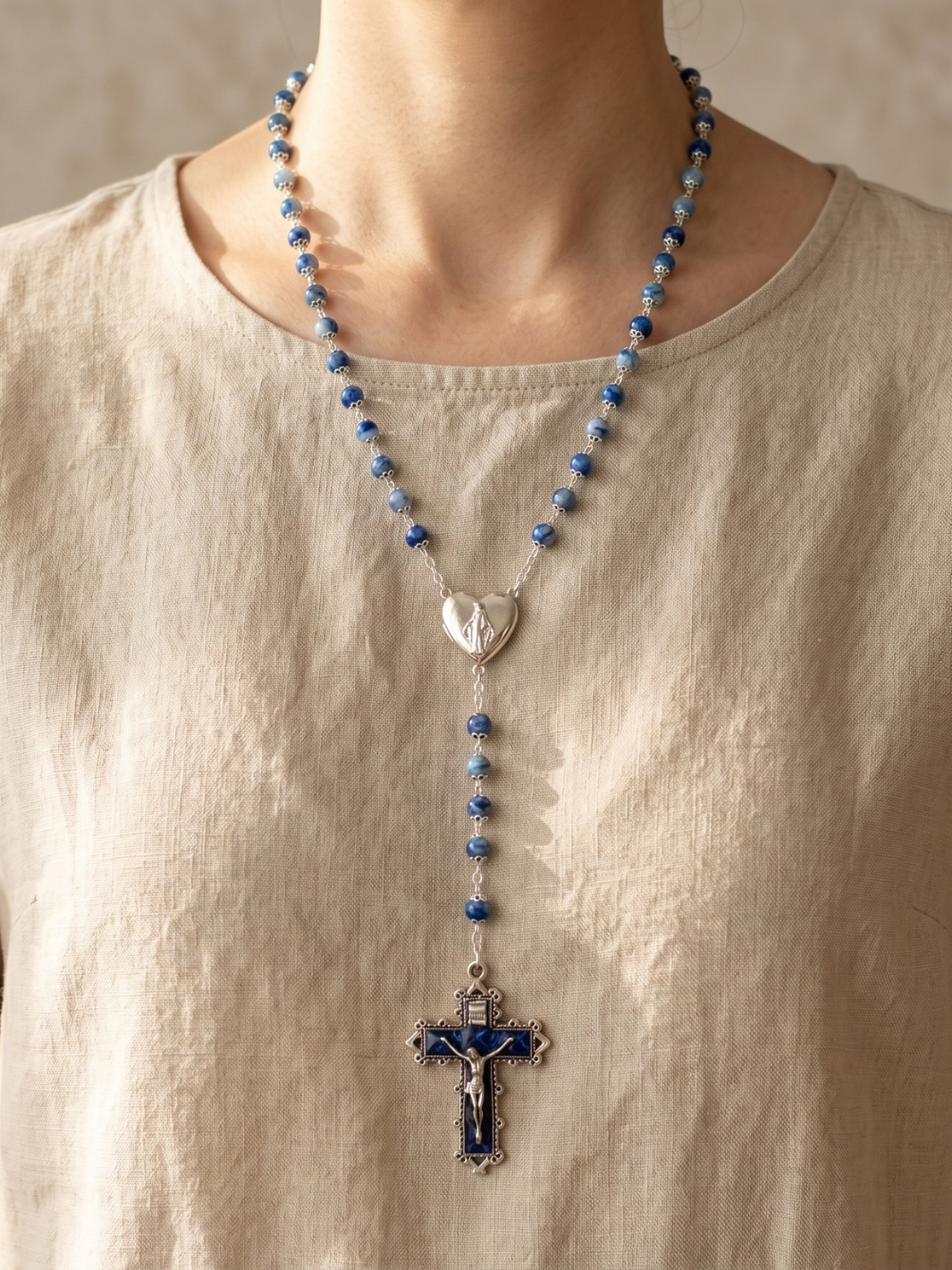 Person wearing a blue beaded necklace with a heart pendant and cross on a beige background