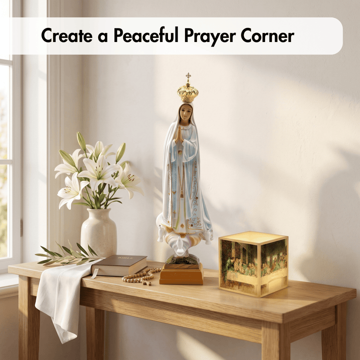 A religious statue of the Virgin Mary with a gold crown stands on a wooden table in a bright room, alongside white lilies in a vase, a book, a cloth, rosary beads, and a small gold box. Text at the top reads, &