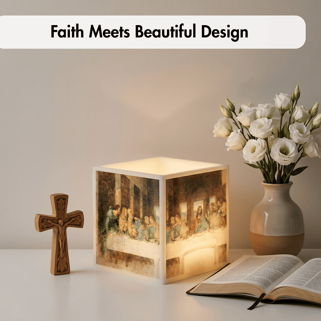 A glowing cube-shaped lamp printed with a religious scene, styled on a tabletop beside a wooden cross, an open book, and a vase of white flowers. Text at the top reads &