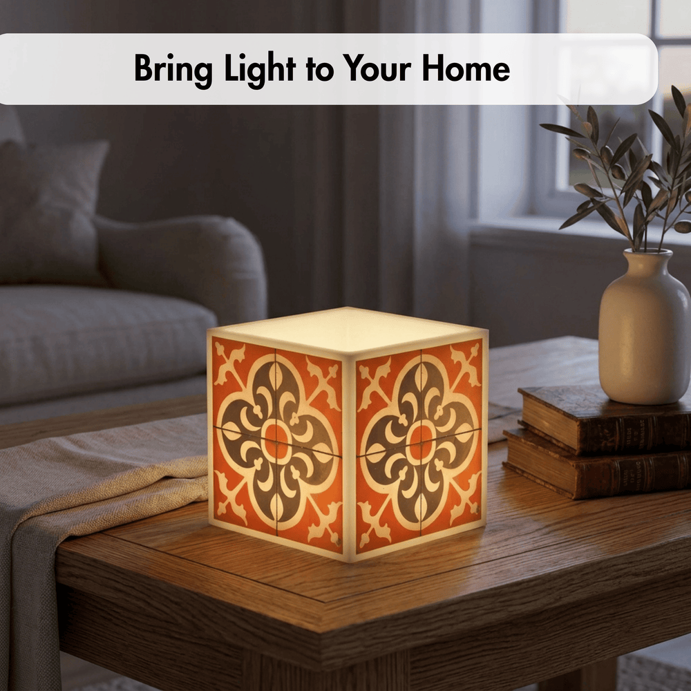 A glowing cube-shaped decorative lamp with a warm orange and cream floral pattern sits on a wooden table in a softly lit living room. A text banner at the top reads, 'Bring Light to Your Home.