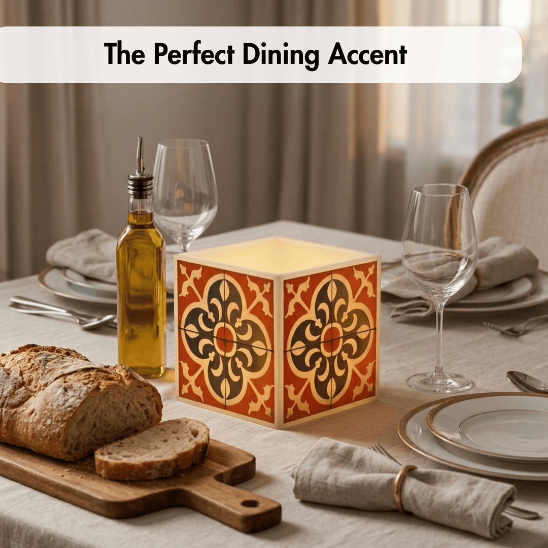 A decorative square table lamp with a warm glowing cream interior and a terracotta orange, black, and gold tile-style geometric pattern on the exterior, styled on a dining table with bread, olive oil, plates, and wine glasses. Text overlay at the top reads &
