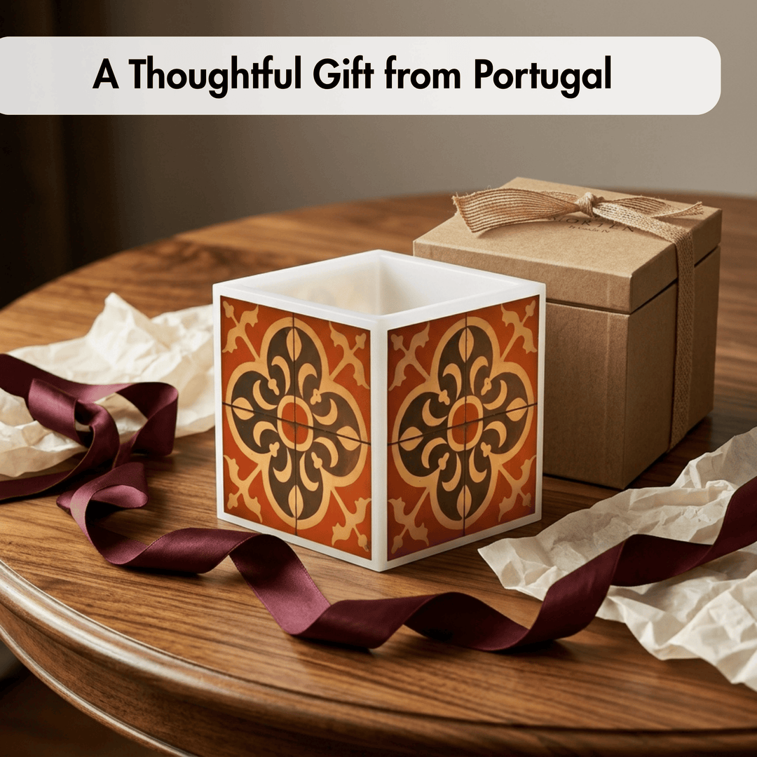 A square gift box with a white interior and orange, brown, and black ornamental tile-style pattern on the outside, displayed on a wooden table with a brown kraft gift box behind it, crumpled tissue paper, and a burgundy ribbon. Text at the top reads &