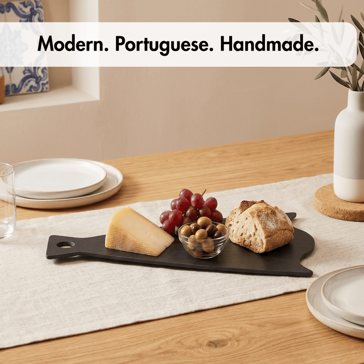 A black rectangular serving board with a handle, styled on a beige table runner with food items including cheese, grapes, olives, and bread on a wooden dining table. White plates and a white vase are in the background.