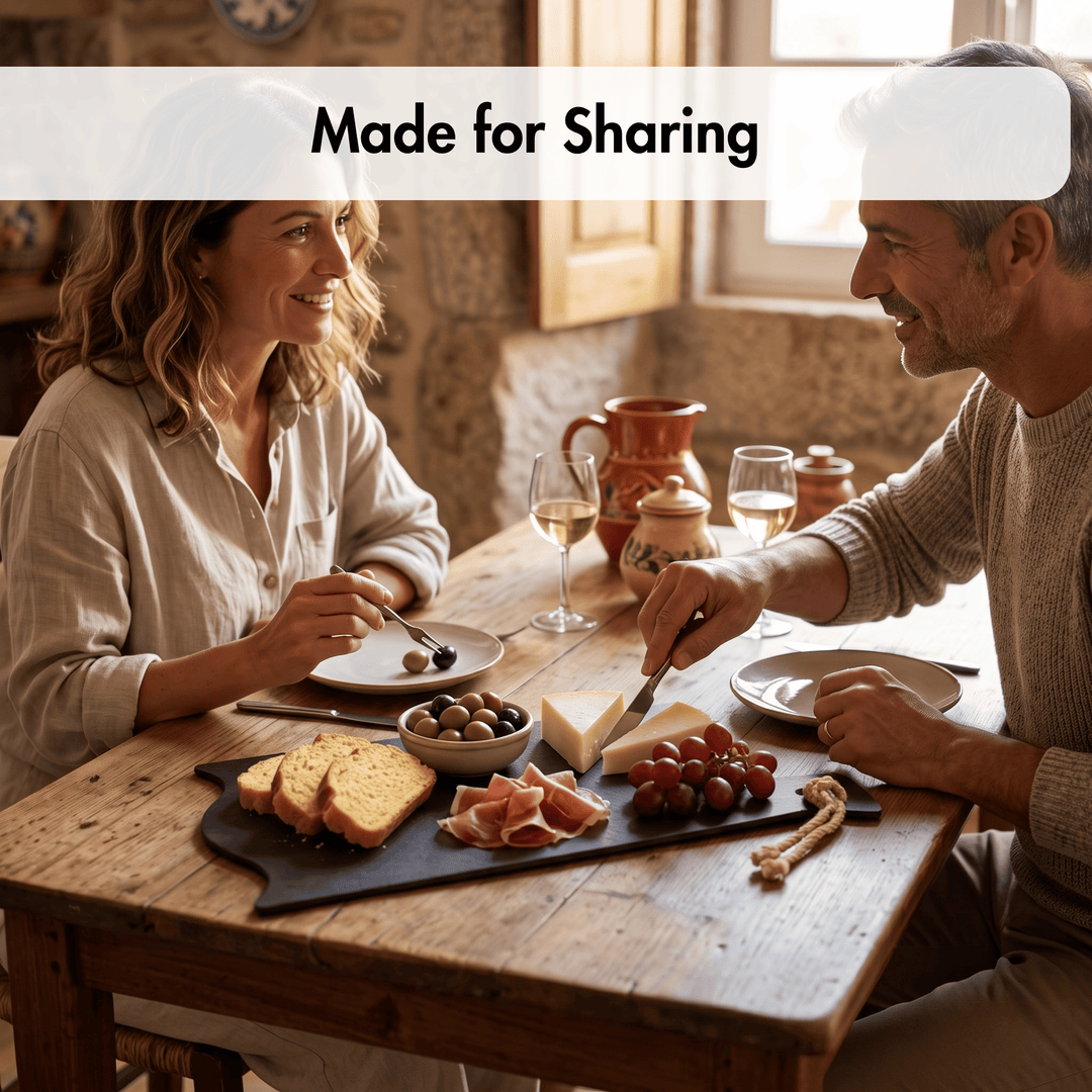 A man and a woman sitting at a rustic wooden table sharing a meal with cheese, bread, grapes, and wine glasses. A white banner at the top reads &