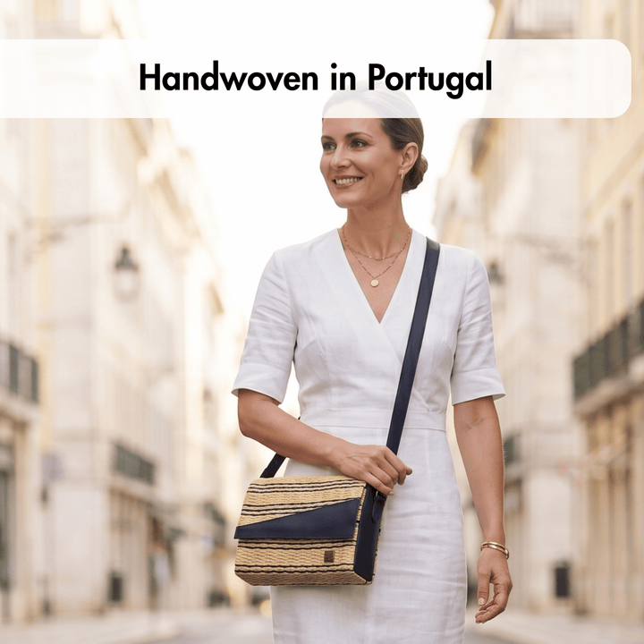 A woman in a white dress stands on a blurred city street in Portugal, wearing a woven shoulder bag with navy blue trim and strap. Text at the top reads &