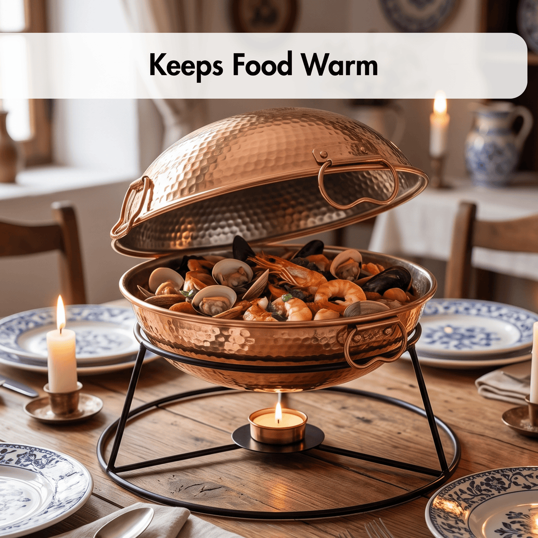 A hammered copper chafing dish with its lid propped open, holding seafood and shellfish on a black metal stand with a small candle underneath. The scene is set on a wooden dining table surrounded by plates, lit candles, and a softly blurred interior background.