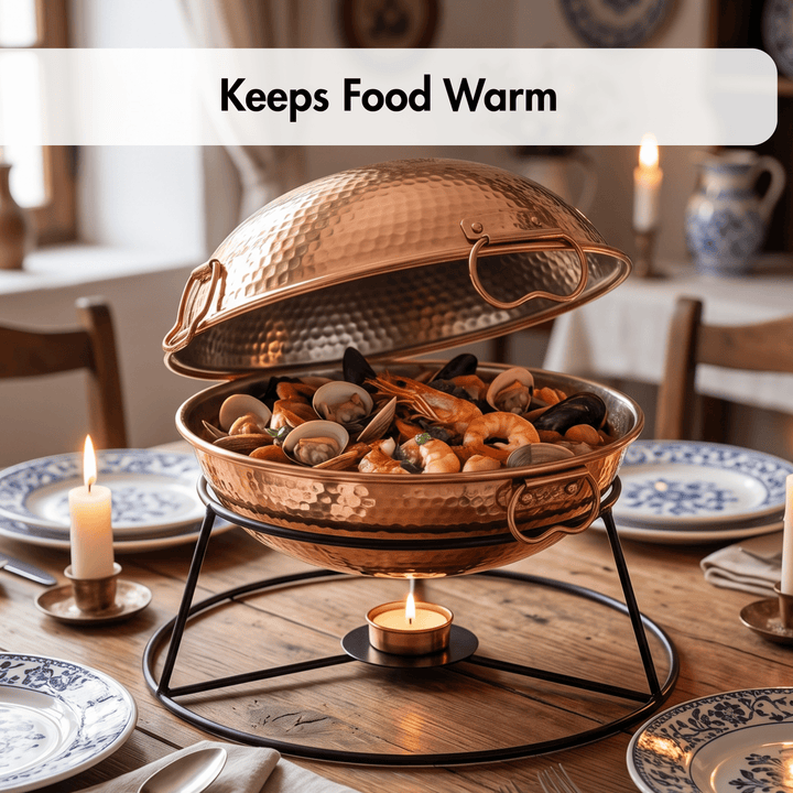 A hammered copper chafing dish with its lid propped open, holding seafood and shellfish on a black metal stand with a small candle underneath. The scene is set on a wooden dining table surrounded by plates, lit candles, and a softly blurred interior background.