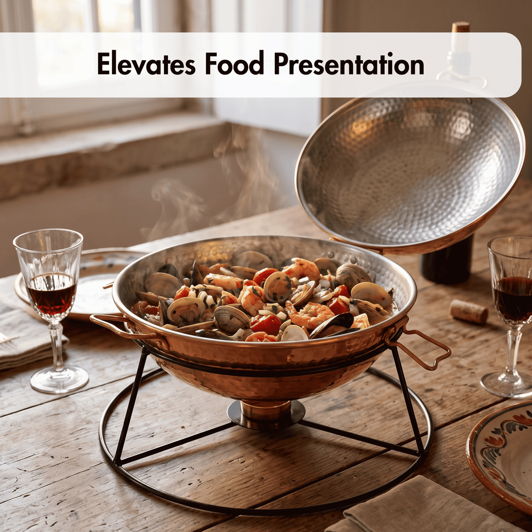 A copper paella pan with a hammered finish sits on a black metal stand on a rustic wooden table, with its lid propped open. Steam rises from the seafood paella inside, and wine glasses are placed on either side.