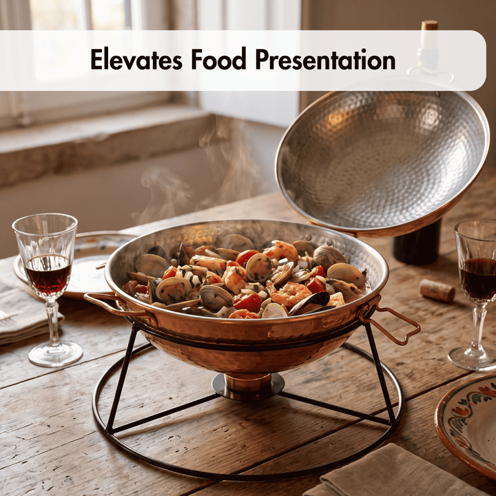 A copper paella pan with a hammered finish sits on a black metal stand on a rustic wooden table, with its lid propped open. Steam rises from the seafood paella inside, and wine glasses are placed on either side.