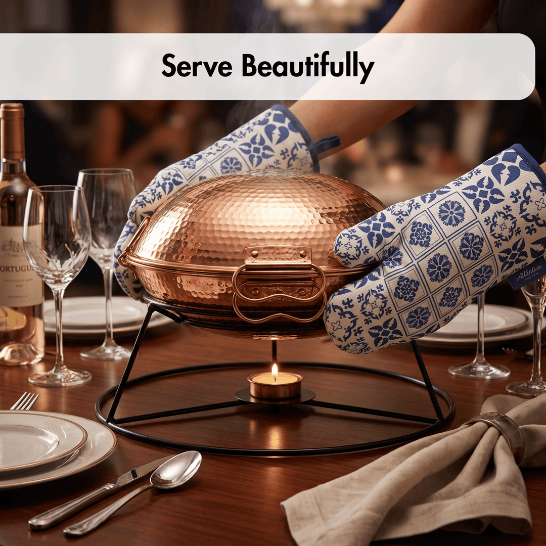 A hammered copper chafing dish sits on a black metal stand on a wooden dining table, held by two hands wearing blue-and-white patterned oven mitts. The scene includes wine glasses, plates, a bottle, and a folded napkin, with the text &