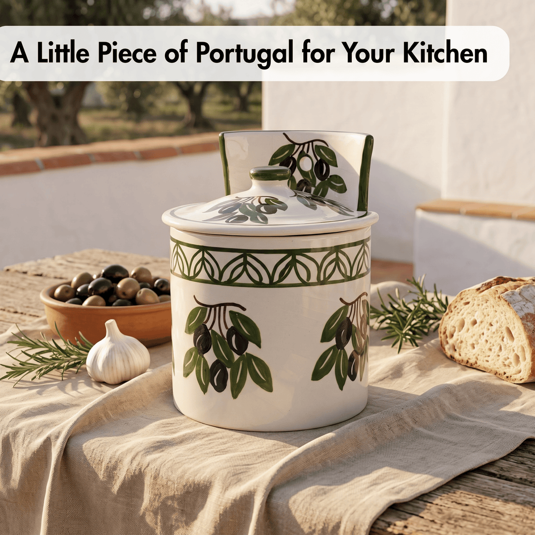 A white ceramic kitchen jar with a lid, decorated with green and black olive branch motifs, set on a beige linen cloth outdoors with bread, garlic, and olives nearby.