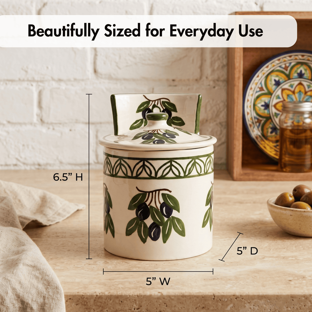 A ceramic canister with a matching lid, decorated with green olive branches and black olives, shown on a kitchen counter with dimension lines indicating 6.5 inches high, 5 inches wide, and 5 inches deep.