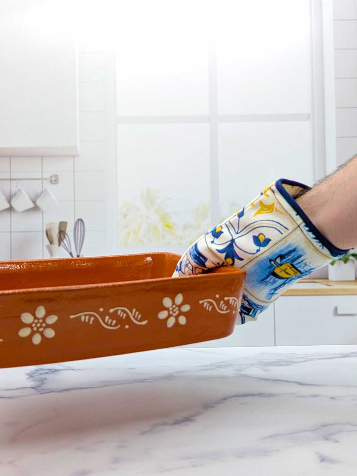 Person wearing a colorful oven mitt holding an orange baking dish with floral patterns in a kitchen.