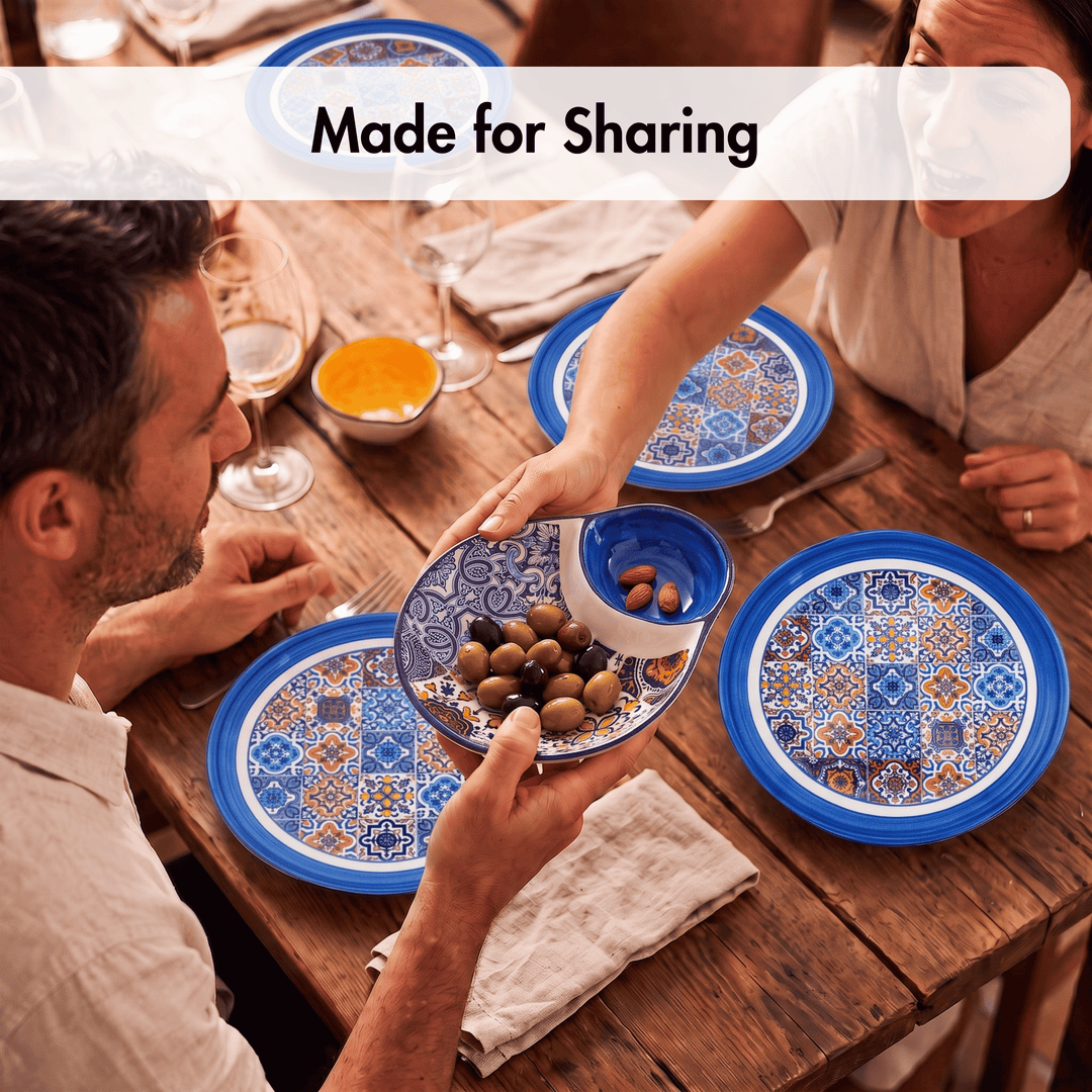A lifestyle shot of a family sharing a meal at a rustic wooden table, featuring a set of blue and white patterned ceramic plates and bowls. A man on the left and a woman on the right are holding serving dishes with olives and bread.