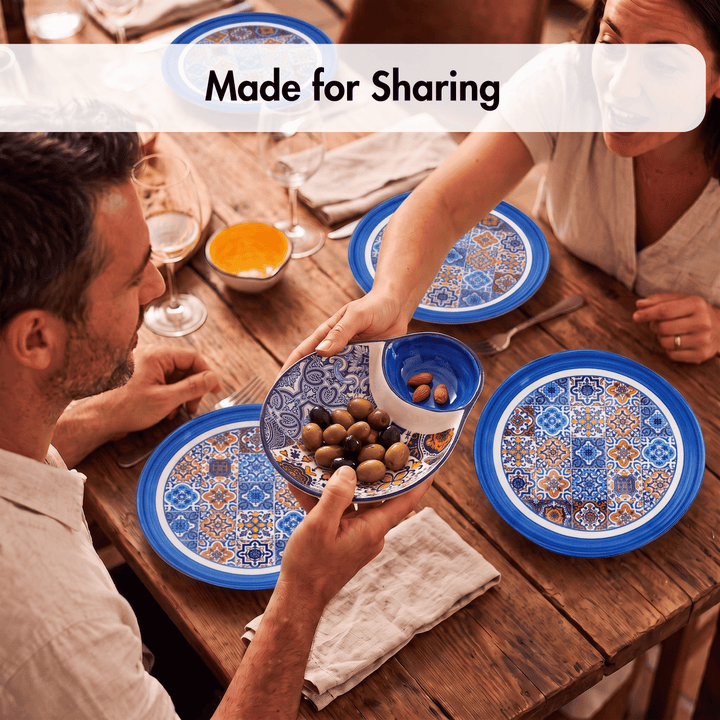 A lifestyle shot of a family sharing a meal at a rustic wooden table, featuring a set of blue and white patterned ceramic plates and bowls. A man on the left and a woman on the right are holding serving dishes with olives and bread.