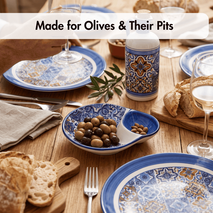 A styled dining table setting featuring a blue and white ceramic olives and pits bowl with a white serving spoon, alongside patterned plates, bread, wine, and a matching patterned bottle. A text banner at the top reads &