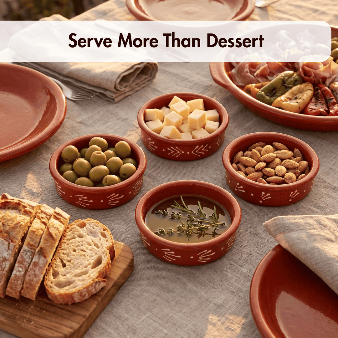 A styled tabletop serving scene with several small terracotta bowls holding olives, cubed cheese, nuts, and a herb oil, alongside bread, plates, and a dish of cured meat. A semi-transparent banner at the top reads “Serve More Than Dessert.