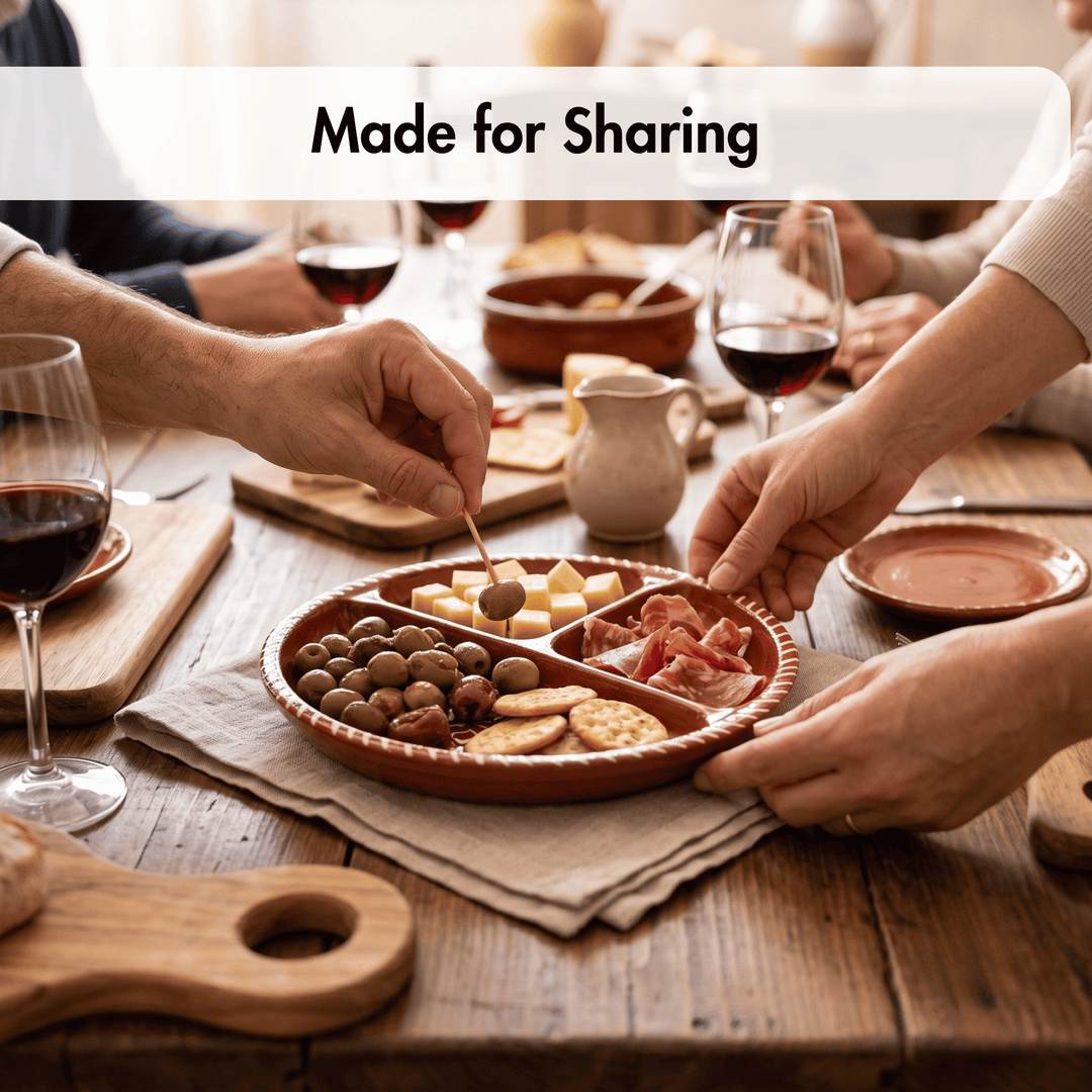 A terracotta clay serving platter with four compartments, filled with crackers, cheese, olives, and cured meat, being shared on a rustic wooden dining table with wine glasses and hands visible. A white banner at the top reads &