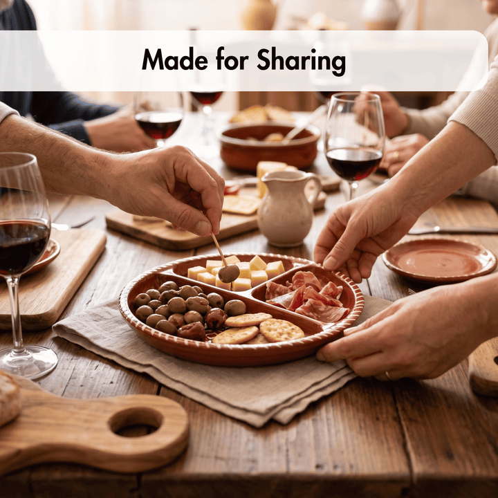 A terracotta clay serving platter with four compartments, filled with crackers, cheese, olives, and cured meat, being shared on a rustic wooden dining table with wine glasses and hands visible. A white banner at the top reads &