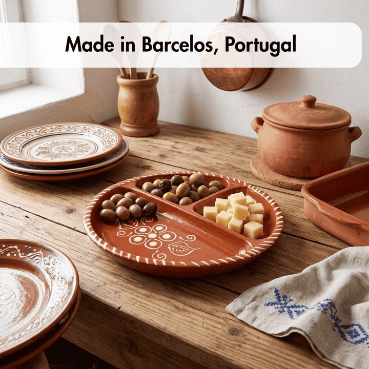 A rustic terracotta clay serving platter with three compartments, styled on a wooden table with olives and cheese cubes. Other matching clay dishes and a lidded pot are visible in a warm, sunlit kitchen setting, with a text overlay reading &