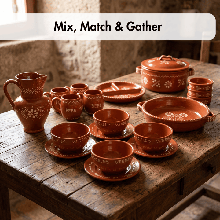 A rustic terracotta clay dinnerware set arranged on a weathered wooden table. The set includes a pitcher, multiple cups and mugs with floral patterns and the words &