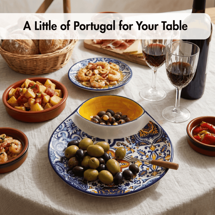 A styled table setting with Portuguese tableware, featuring a large blue and yellow patterned platter with olives, a yellow-rimmed bowl, smaller terracotta bowls, bread, and two glasses of red wine. A text overlay at the top reads &