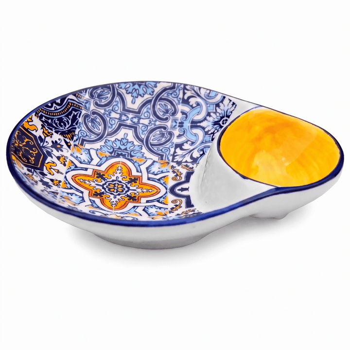 A ceramic Spanish-style dip bowl with a white base, blue and orange floral patterns on the interior, and a glossy yellow-orange glazed recess for the dip, shown on a white background.
