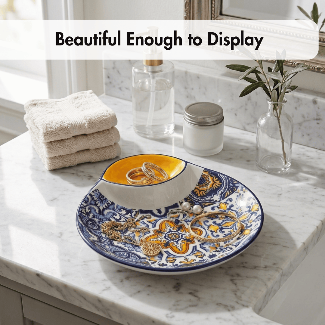 A decorative oval jewelry display tray with a blue and gold floral pattern, holding a small white bowl with a yellow interior and rings, set on a white marble bathroom counter with folded towels, glass bottles, and a small vase in the background. Text overlay at the top reads &