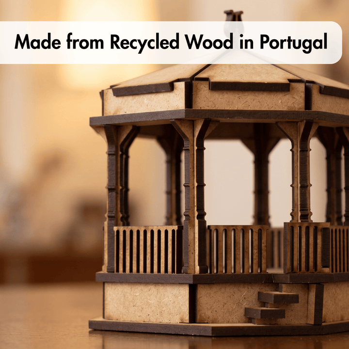 A small wooden gazebo or pavilion model made from recycled wood, shown on a tabletop with a warm blurred background. A white text banner at the top reads &