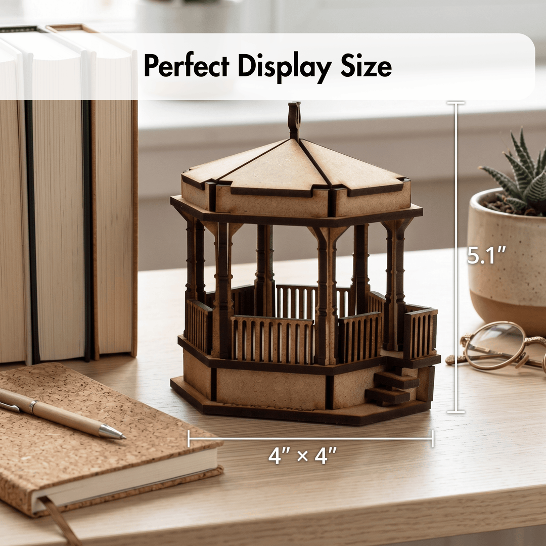 A wooden gazebo model displayed on a light wood desk with measurement annotations showing 4" x 4" base and 5.1" height, alongside books, a notebook, pen, and small potted plant.