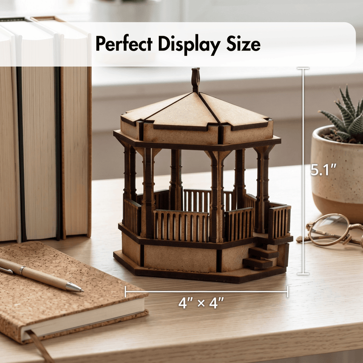 A wooden gazebo model displayed on a light wood desk with measurement annotations showing 4" x 4" base and 5.1" height, alongside books, a notebook, pen, and small potted plant.