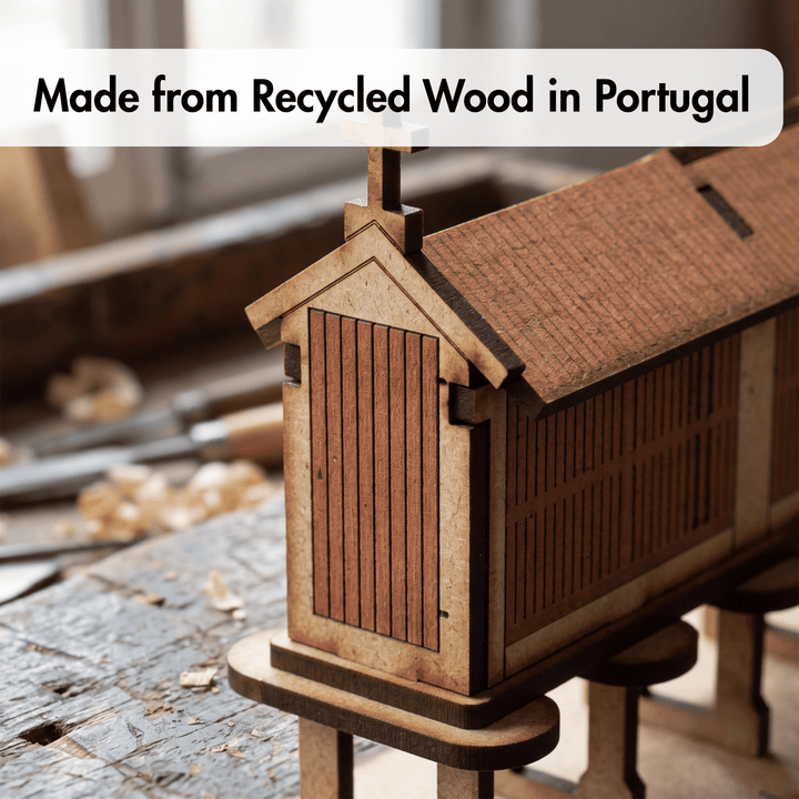 A small wooden house model or miniature cabin made from recycled wood, shown on a workbench with blurred workshop tools in the background. A text banner at the top reads &