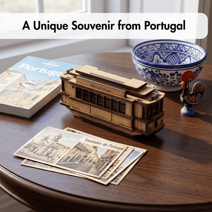 A wooden model of a Lisbon street car on a dark wooden table, surrounded by travel-themed items including a book, postcards, a blue and white ceramic bowl, and a small rooster figurine. A text banner at the top reads &