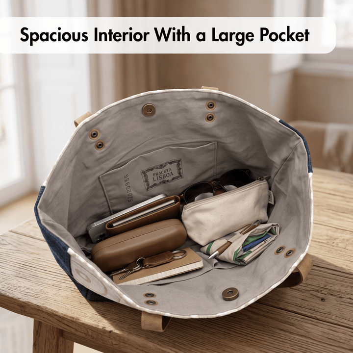 An open tote bag shown from the inside, placed on a wooden table in a softly lit room. The bag has a light gray interior with brass-tone snap closures and a front pocket, and it is filled with small accessories like sunglasses, a wallet, and a pouch.