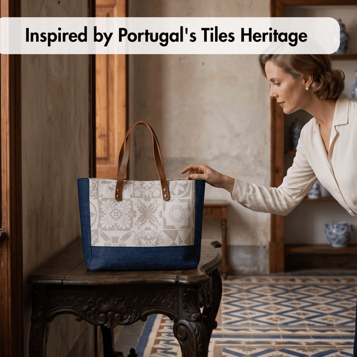A woman in a cream blouse leans toward a tote bag placed on a carved wooden table in an indoor room. The bag has a navy blue base and top handles, with a large cream panel featuring a geometric tile-inspired pattern.